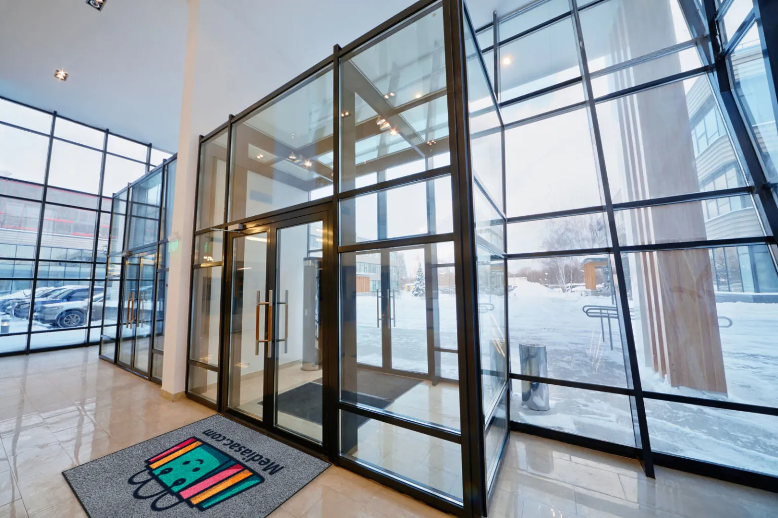 Entrée vitrée moderne avec un grand tapis personnalisé portant un logo coloré et l’adresse d’un site web.